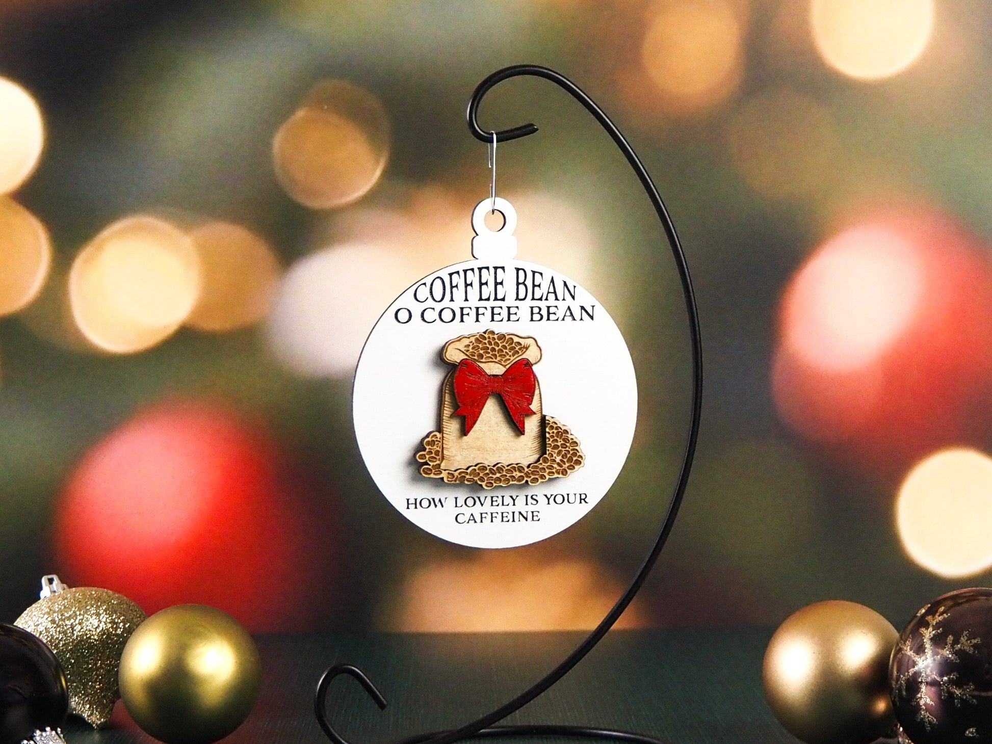 Round white ornament hanging from a black metal stand, featuring the phrase "Coffee Bean O Coffee Bean" at the top and "How lovely is your caffeine" at the bottom. The design includes a burlap sack filled with coffee beans, decorated with a red bow. Holiday ornaments are placed nearby.