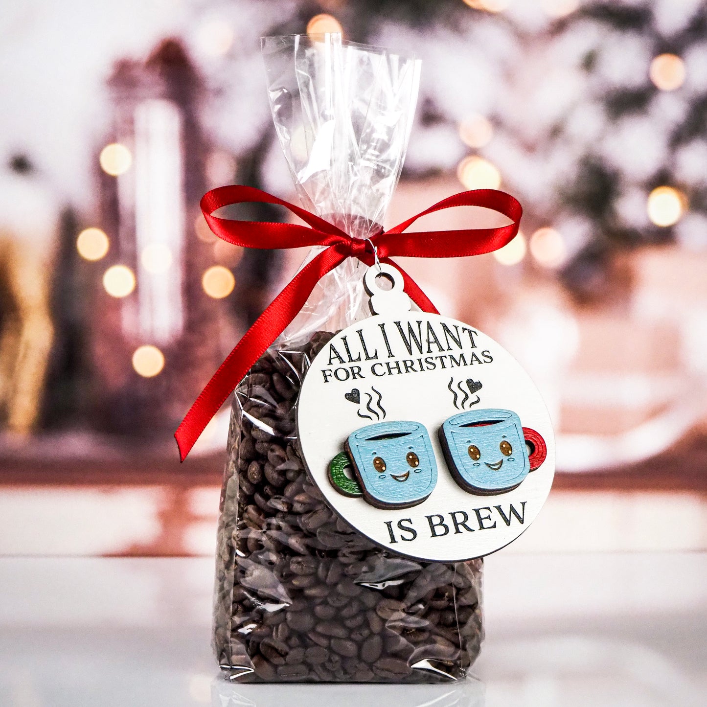 Clear bag of coffee beans tied with a red ribbon, featuring a round white ornament attached. The ornament has black text that reads "All I want for Christmas is brew," with two smiling blue mugs, one with a green handle and one with a red handle, with steam rising above them.