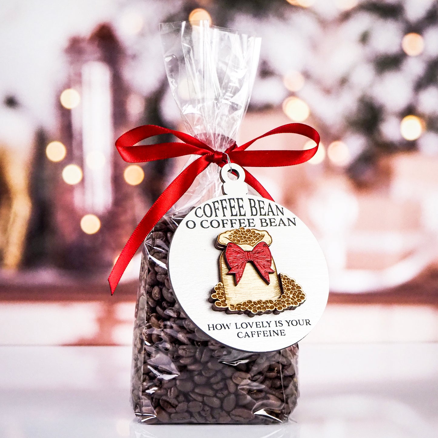 Clear bag of coffee beans tied with a red ribbon, featuring a round white ornament. The white ornament has black text "Coffee Bean O Coffee Bean" at the top and "How lovely is your caffeine" at the bottom. The design includes a burlap sack filled with coffee beans and a red bow.