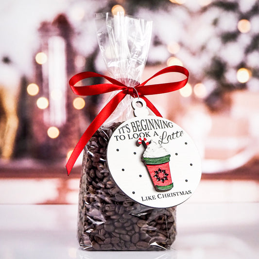 White round ornament with the text "It's Beginning to Look a Latte Like Christmas" featuring a raised red coffee cup design with whipped cream and a candy cane. The ornament is tied to a bag of coffee beans with a red ribbon, set against a holiday-themed background with blurred lights.