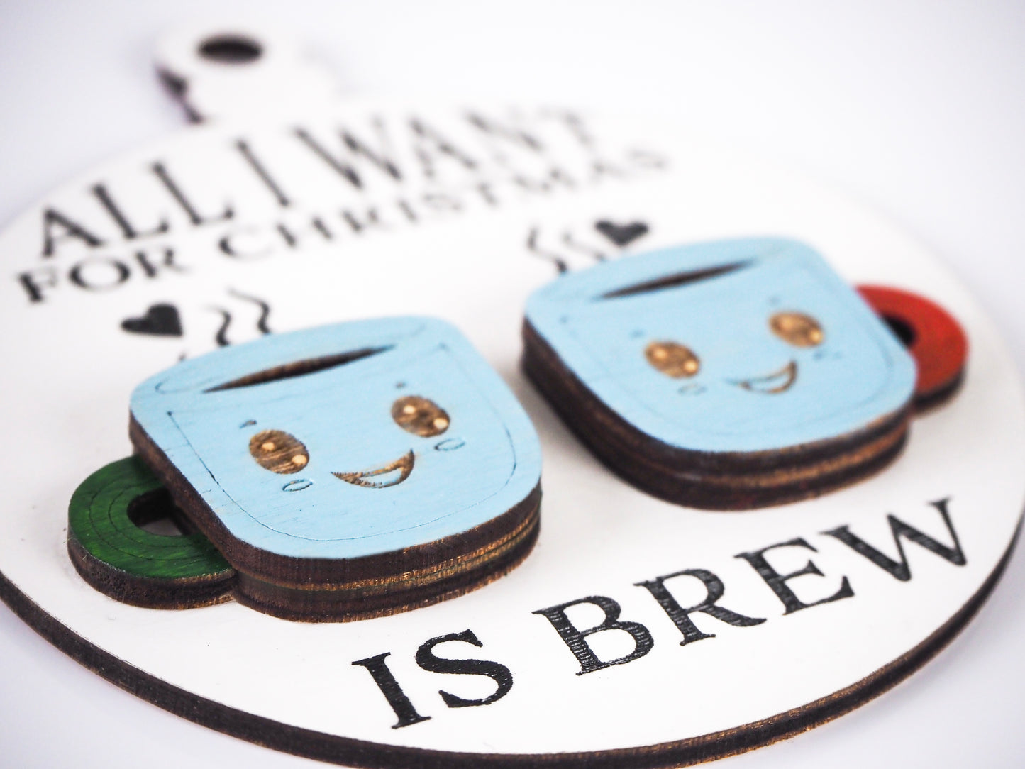 Close-up of a round white ornament with black text that reads "All I want for Christmas is brew." The ornament features two smiling blue mugs with brown eyes, one with a green handle and the other with a red handle, with steam rising from the mugs.