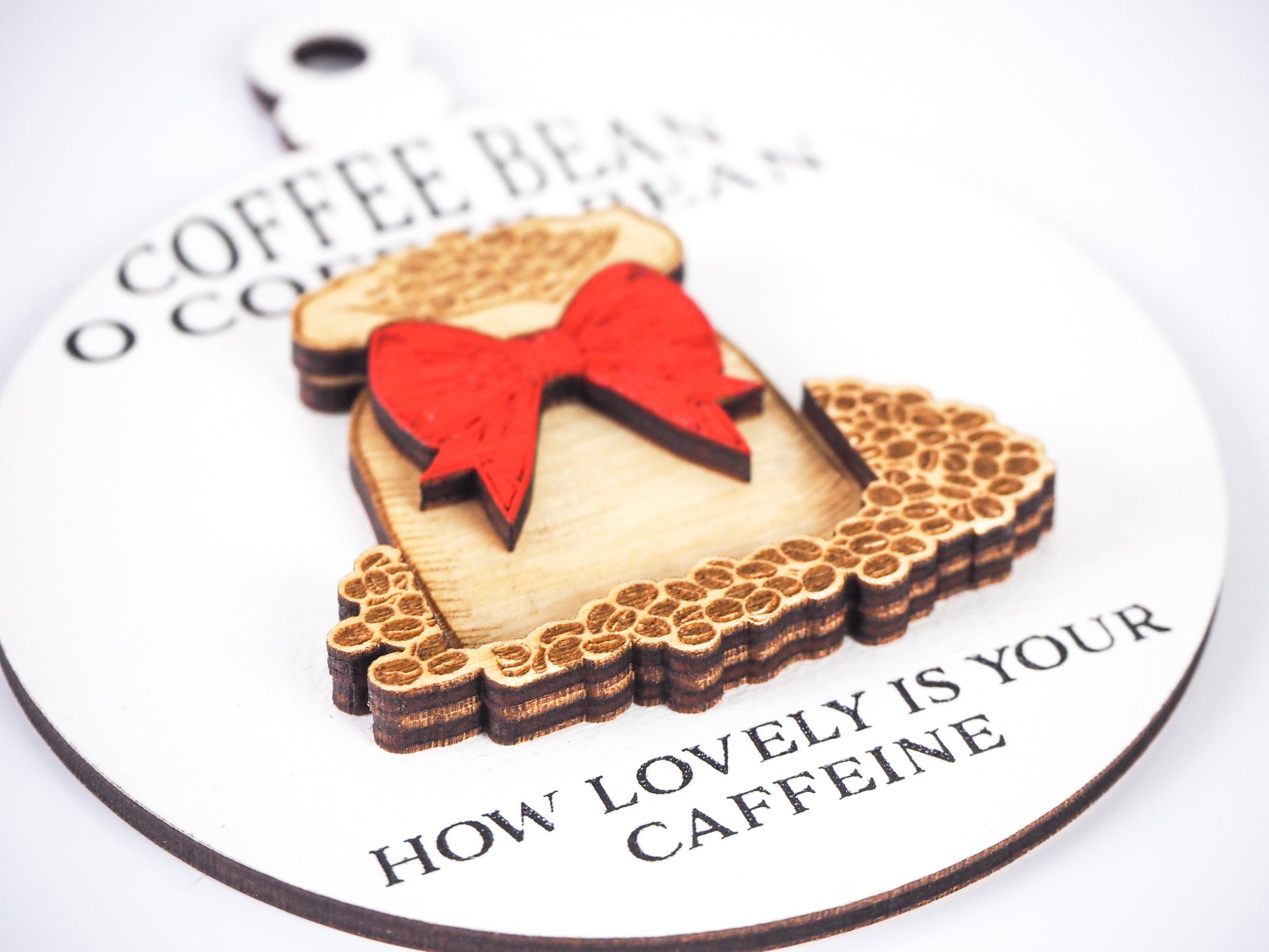 Close-up of a white round ornament featuring the text in black "Coffee Bean O Coffee Bean" at the top and "How lovely is your caffeine" at the bottom. The design includes a raised burlap sack with a red bow and intricate wood detailing of coffee beans.