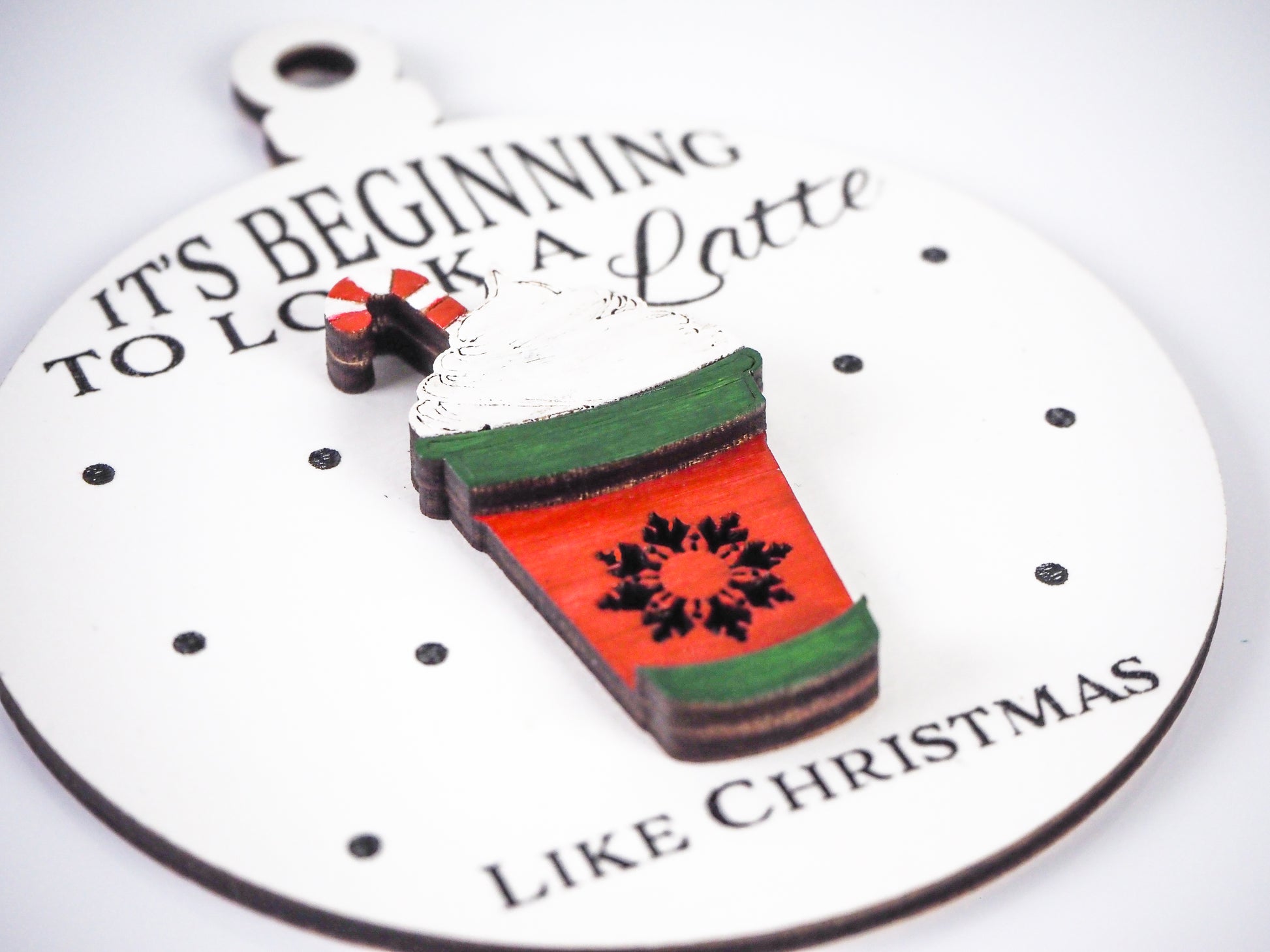 Close-up of a white ornament with the raised design of a red coffee cup, green accents, whipped cream, and a candy cane on top. The text on the ornament reads, "It's Beginning to Look a Latte Like Christmas" with small black dots as snowflakes on a white background.