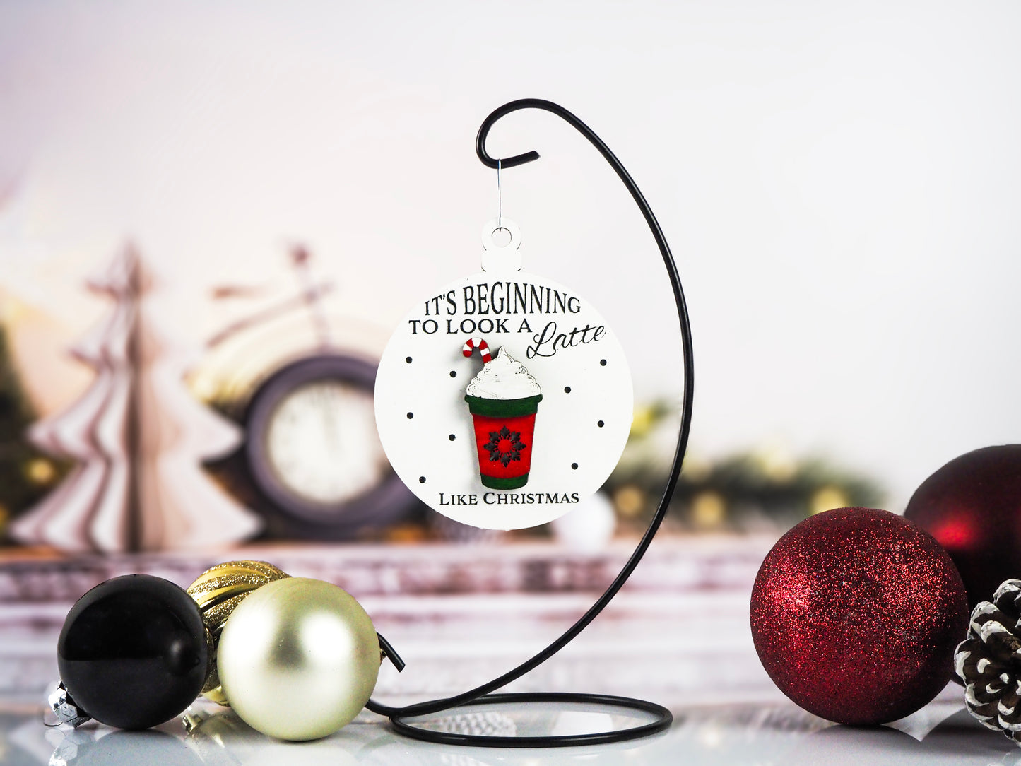 White round ornament with the text "It's Beginning to Look a Latte Like Christmas" and a raised red coffee cup design featuring whipped cream, a candy cane, and snowflakes. The background includes festive holiday décor and ornaments in black, gold, and red.