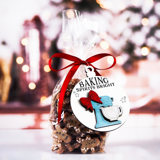 Bag of gingerbread cookies tied with a red ribbon and decorated with a round ornament featuring a raised blue mixer design with a red bow. The text on the white ornament reads, "Baking Spirits Bright," with small stars around the design.