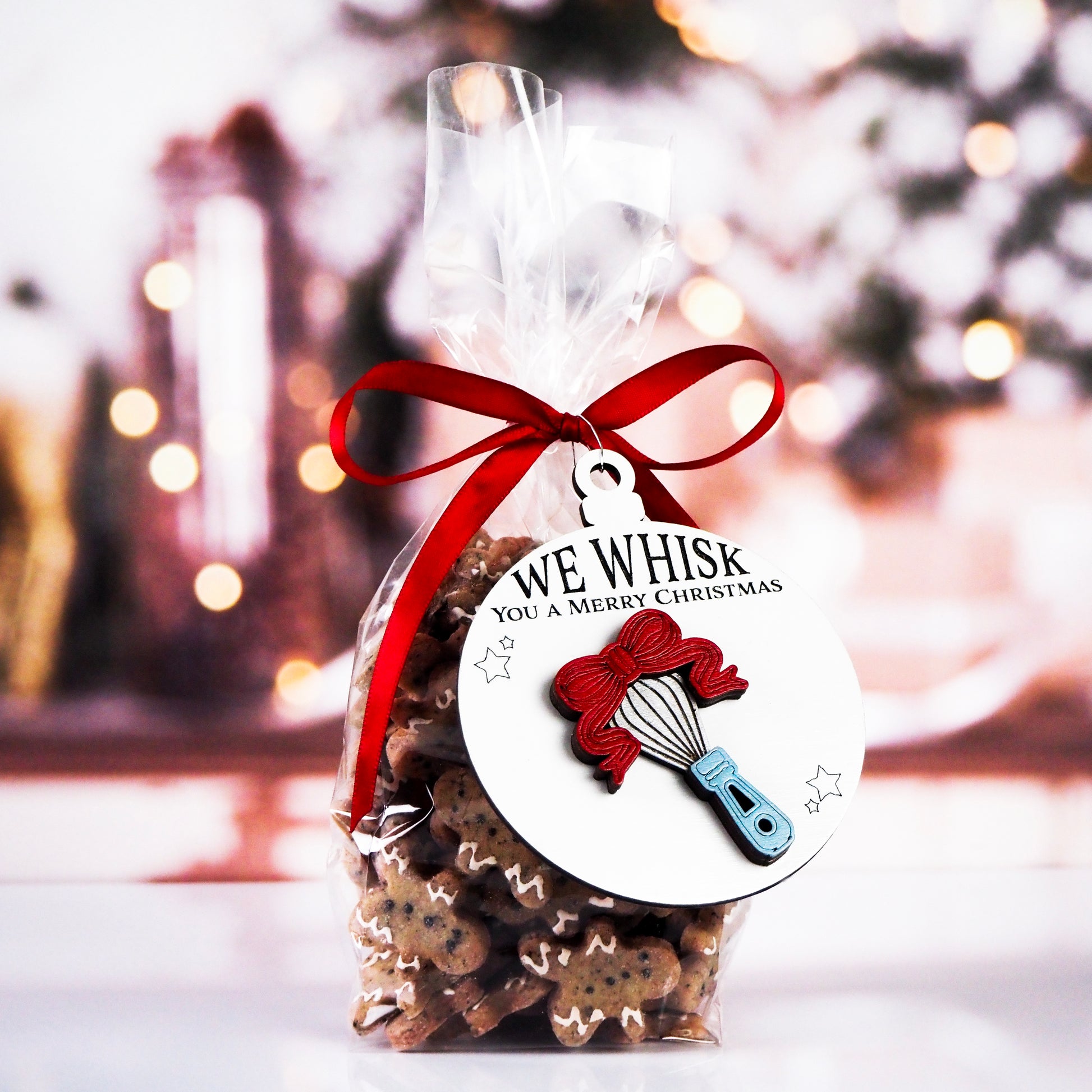 Clear bag of gingerbread cookies tied with a red ribbon and a round ornament featuring a raised whisk design with a red bow. The white ornament reads, "We Whisk You a Merry Christmas" with small stars around the whisk.