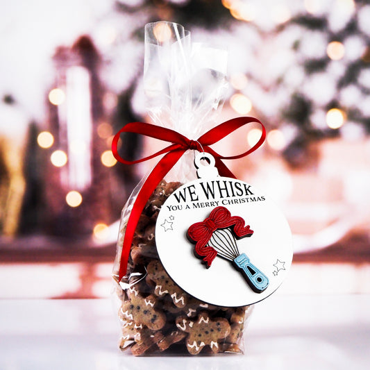 Clear bag of gingerbread cookies tied with a red ribbon and a round ornament featuring a raised whisk design with a red bow. The white ornament reads, "We Whisk You a Merry Christmas" with small stars around the whisk.