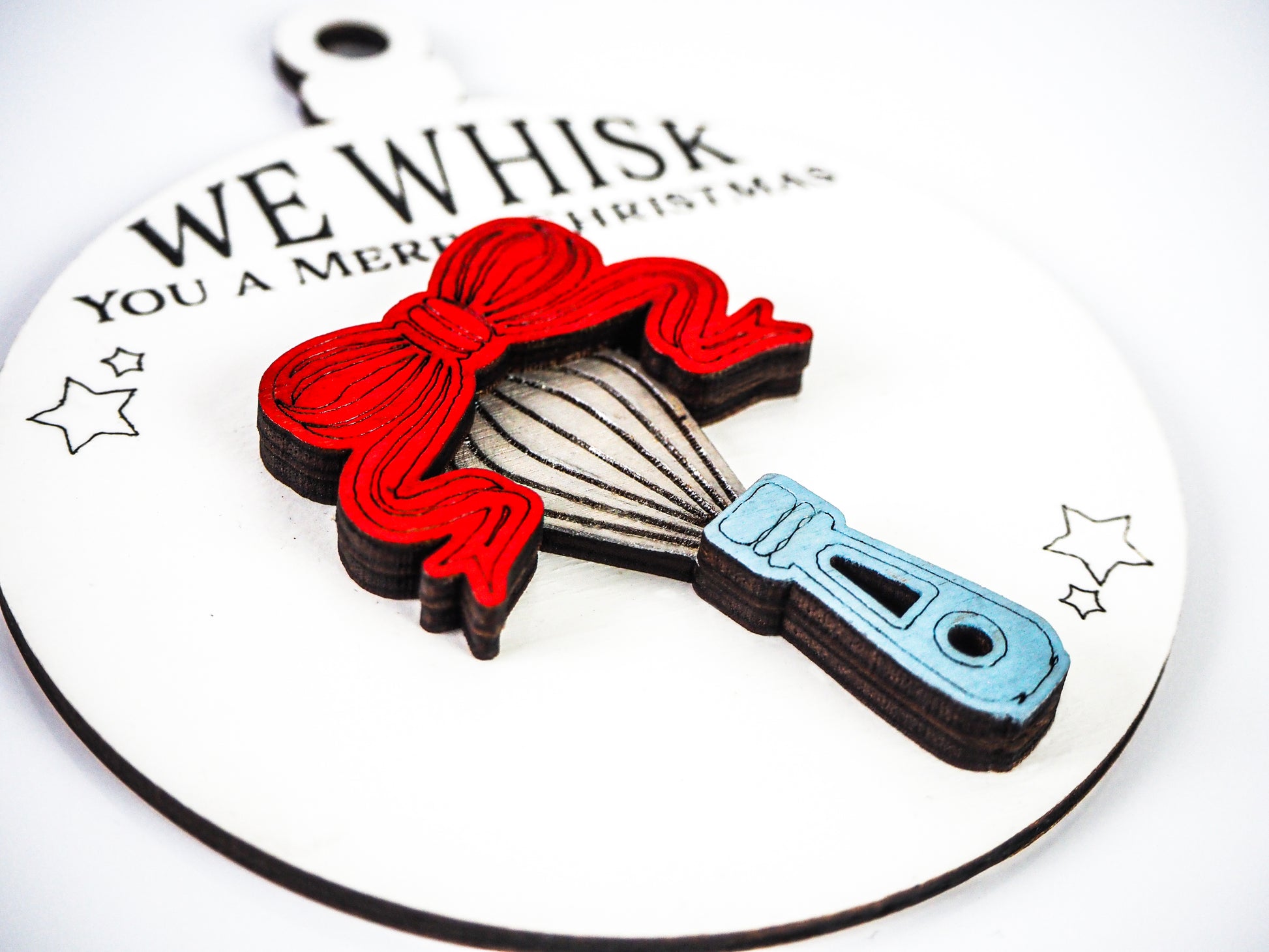 Close-up of a Christmas ornament featuring a raised whisk design with a red bow and a light blue handle on a white background. The text on the ornament reads "We Whisk You a Merry Christmas," with small stars drawn around the whisk.