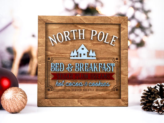 Wooden sign with the text "North Pole Bed & Breakfast" featuring a light blue house and tree design. The sign reads "Sugar Plum Dreams, Hot Cocoa & Cookies Served Nightly" in blue, white, and red text, with holiday ornaments in the background.