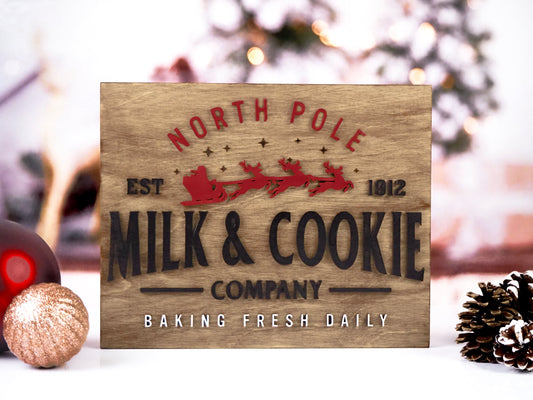 North Pole Milk & Cookie Company sign featuring red Santa sleigh with reindeer and black text on a wooden background. Text reads "Baking Fresh Daily" in white, creating a rustic holiday look.