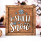 Wooden framed "I Smell Snow" sign with 3D white text and raised blue snowflake accents. The sign is displayed against a cozy, festive background with Christmas ornaments and pinecones.