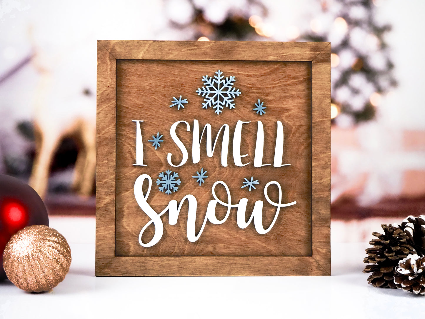 Wooden framed "I Smell Snow" sign with 3D white text and raised blue snowflake accents. The sign is displayed against a cozy, festive background with Christmas ornaments and pinecones.