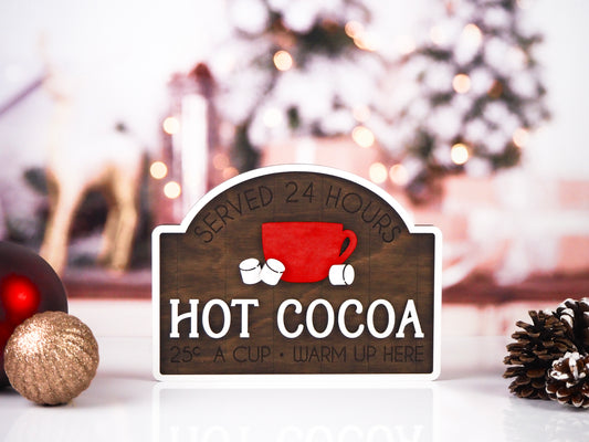 Rustic "Hot Cocoa" sign with a red cup and two marshmallows. The sign reads "Served 24 Hours, Hot Cocoa 25¢ a cup, Warm Up Here" with a cozy winter background, festive ornaments, and pinecones.