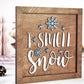 Wooden framed "I Smell Snow" sign with raised white lettering and blue snowflakes. The sign is placed in a festive setting with pinecones and decorative Christmas trees in the background.