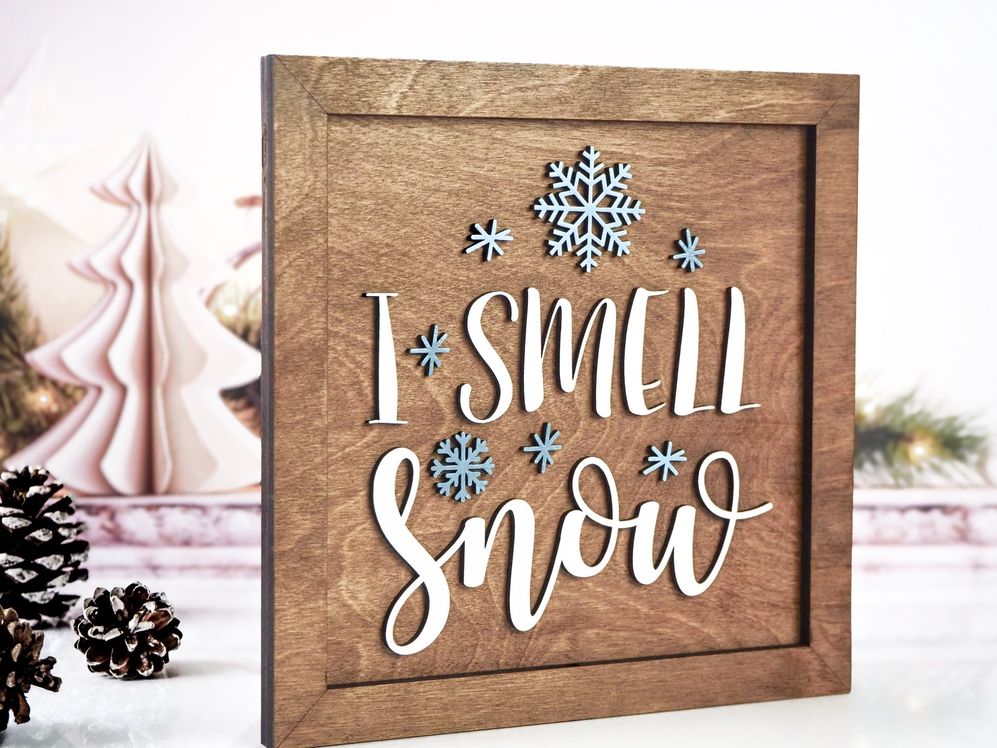 Wooden framed "I Smell Snow" sign with raised white lettering and blue snowflakes. The sign is placed in a festive setting with pinecones and decorative Christmas trees in the background.