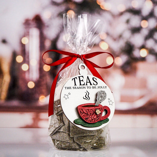 A festive tea gift featuring a wooden ornament with a red teacup design and the phrase "Teas The Season To Be Jolly." The ornament is attached to a clear bag filled with tea bags, tied with a red ribbon, with a blurred holiday background.