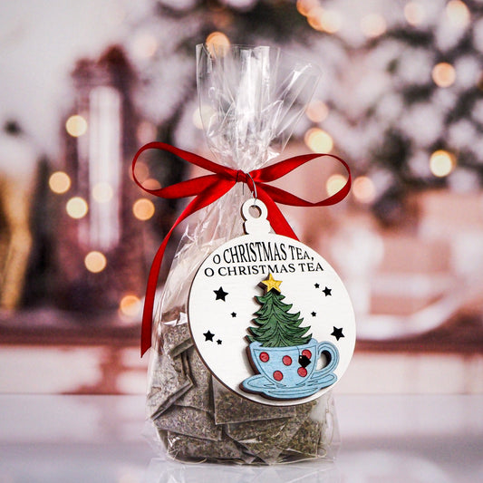 Christmas ornament featuring a whimsical design of a Christmas tree inside a polka-dotted teacup with a star topper. The text on the ornament reads “O Christmas Tea, O Christmas Tea” against a white background with black stars. Tied to a gift bag with red ribbon.