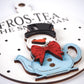 Close-up view of a snowman ornament shaped like a teapot, featuring a black top hat, red bow tie, and blue teapot base. The text "Fros-Tea the Snowman" is visible on the white background with black polka dot accents.