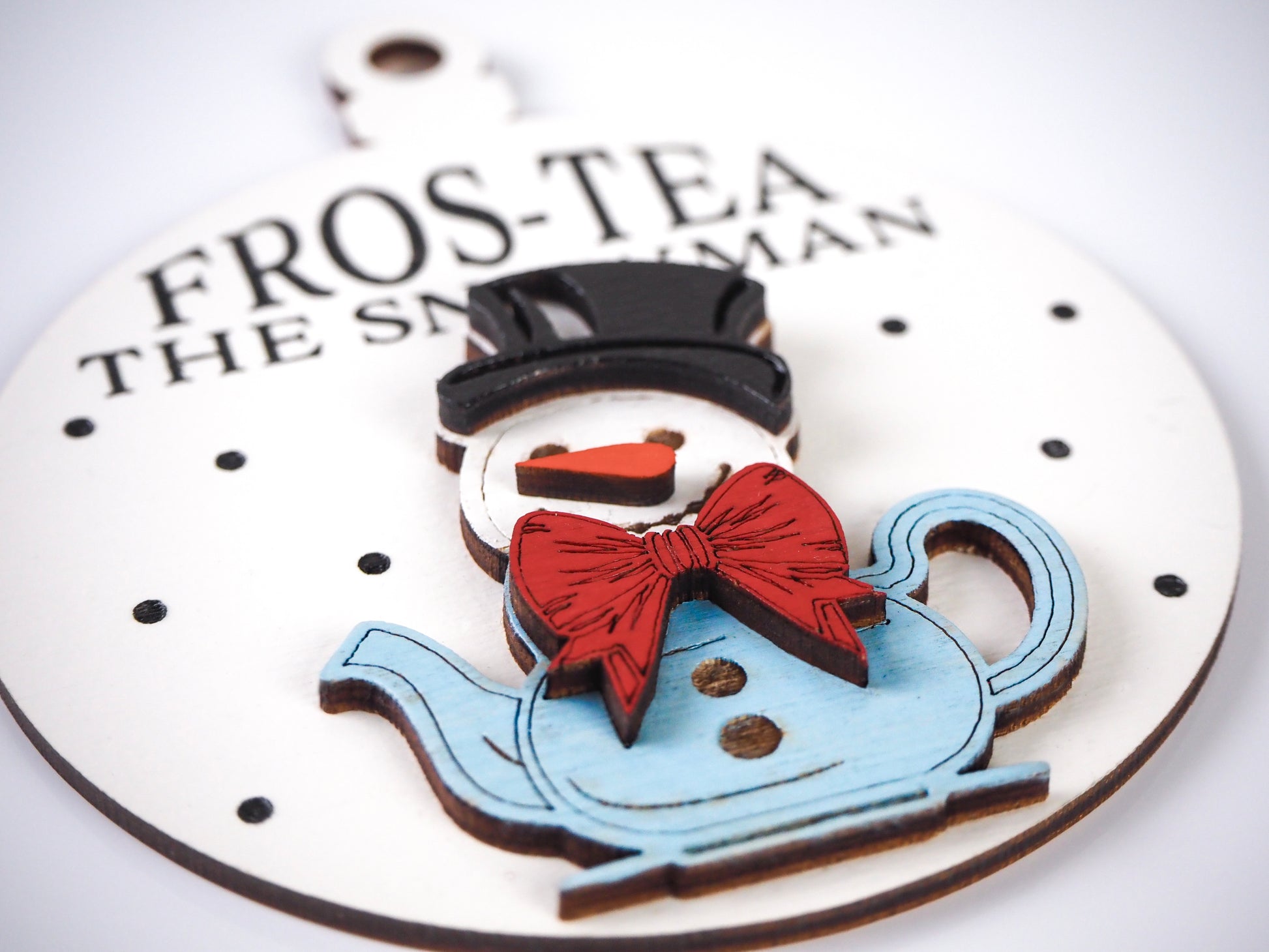 Close-up view of a snowman ornament shaped like a teapot, featuring a black top hat, red bow tie, and blue teapot base. The text "Fros-Tea the Snowman" is visible on the white background with black polka dot accents.