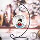 Christmas ornament featuring a snowman in a blue teapot, wearing a black top hat and a red bow tie. The text reads "Fros-Tea the Snowman" on a white background with dotted accents. The ornament is hanging on a black display stand with festive décor in the background.