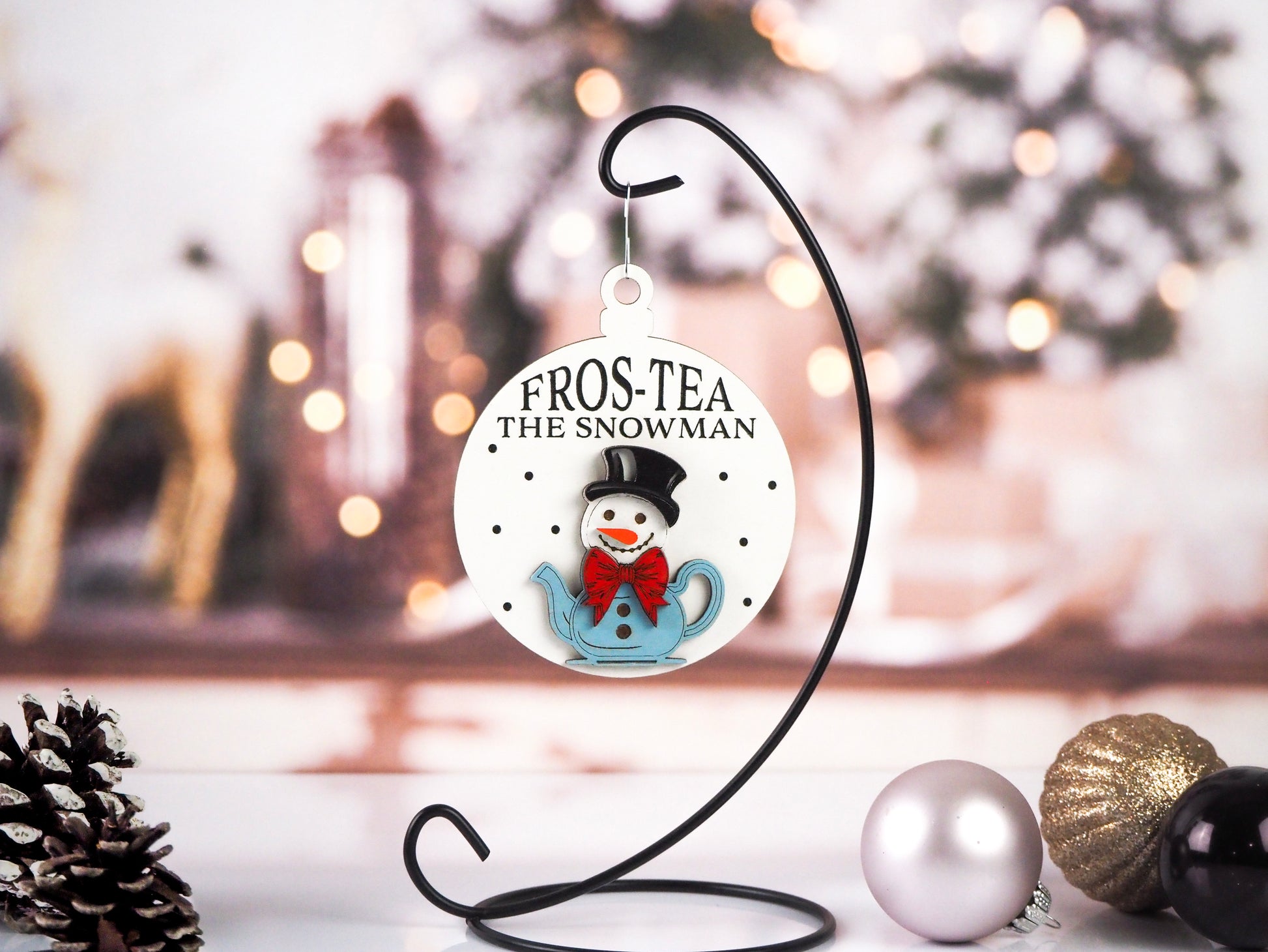 Christmas ornament featuring a snowman in a blue teapot, wearing a black top hat and a red bow tie. The text reads "Fros-Tea the Snowman" on a white background with dotted accents. The ornament is hanging on a black display stand with festive décor in the background.