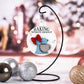 A Christmas ornament featuring a vintage blue stand mixer with a red bow, hanging from a black metal stand. The text "Baking Spirits Bright" is written above the mixer on a white background, with festive holiday ornaments in the foreground.