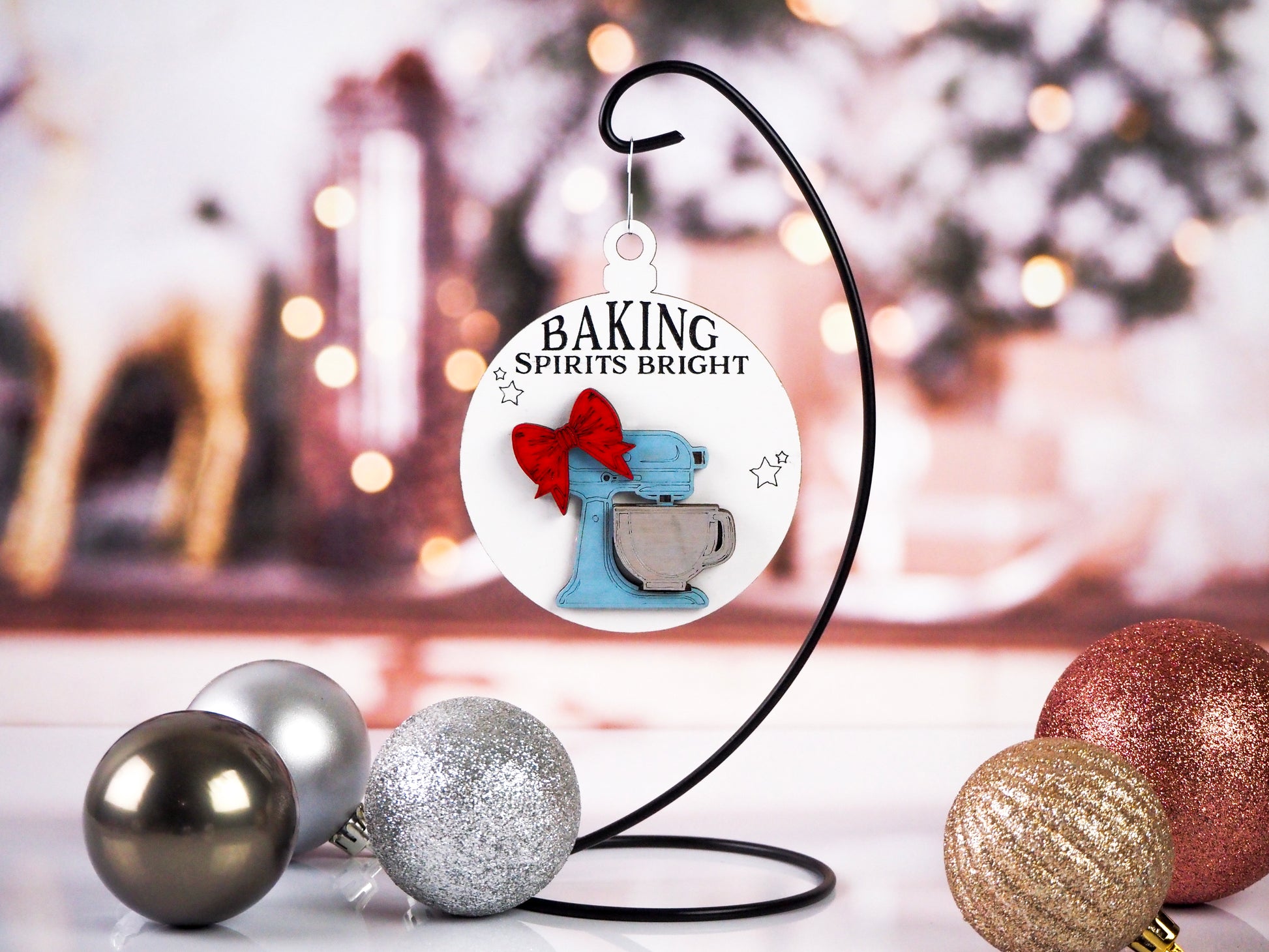 A Christmas ornament featuring a vintage blue stand mixer with a red bow, hanging from a black metal stand. The text "Baking Spirits Bright" is written above the mixer on a white background, with festive holiday ornaments in the foreground.