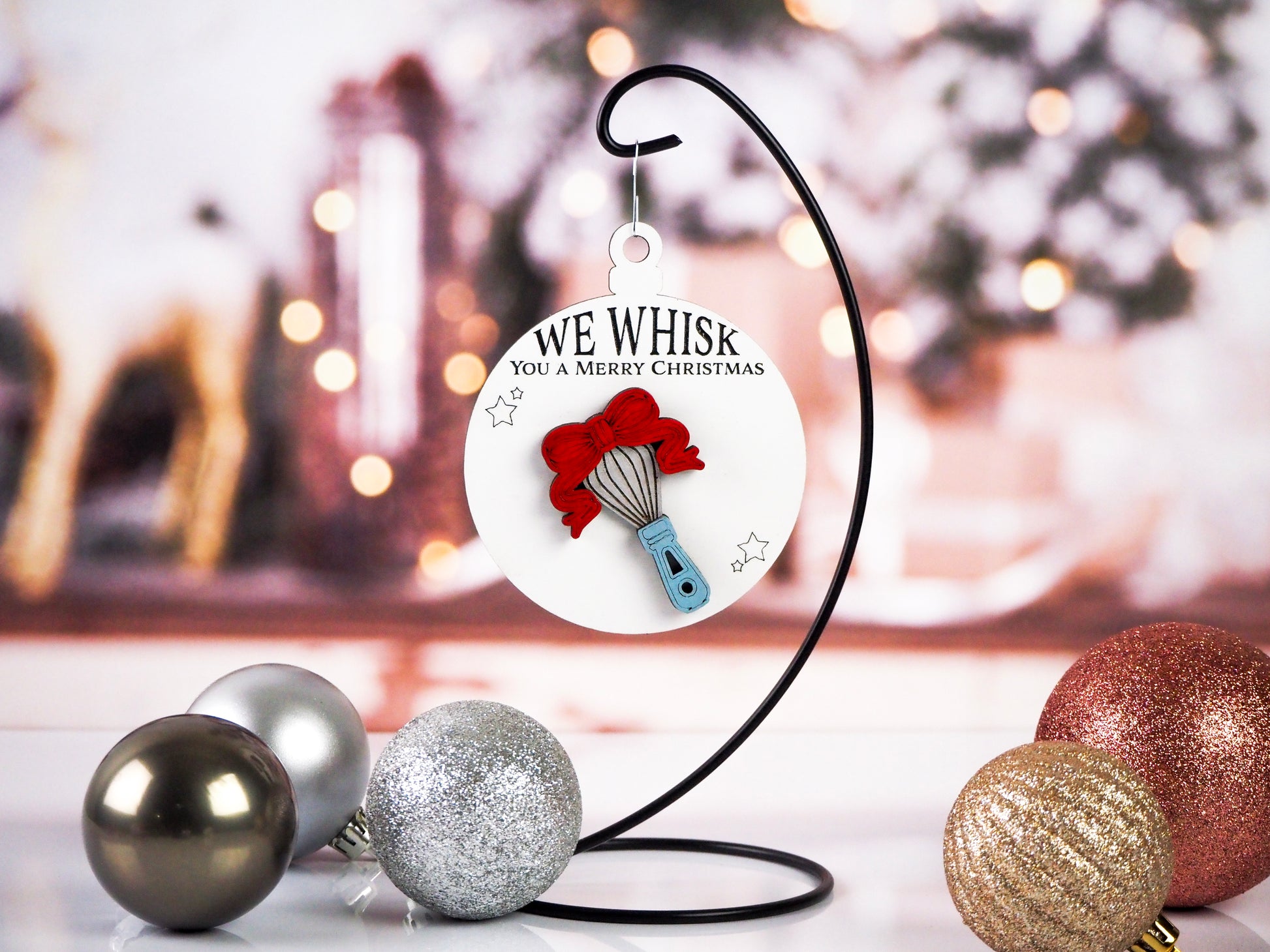 A Christmas ornament featuring a whisk with a red bow on a white background, hanging from a black metal stand. The text reads "We Whisk You a Merry Christmas" above the whisk, with holiday ornaments in the foreground.