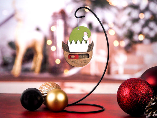 Wooden Christmas ornament shaped like an elf hat with a slider, showing options for "Naughty" or "Nice." The ornament is hung on a black display hook, surrounded by red, gold, and black baubles, set against a festive background.