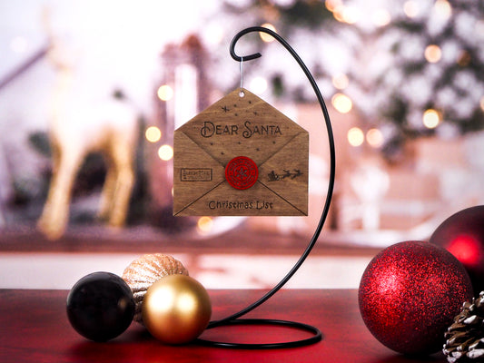 3D wooden envelope ornament addressed to Santa with engraved details, red wax seal, and "Christmas List" text, displayed hanging on a stand with festive background decor. It can hold a small list.