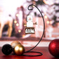 Hanging ornament shaped like a s’more, featuring a marshmallow snowman with twig arms, stacked on a graham cracker base with melted chocolate details. Displayed on a stand with festive background decor.