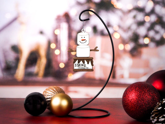 Hanging ornament shaped like a s’more, featuring a marshmallow snowman with twig arms, stacked on a graham cracker base with melted chocolate details. Displayed on a stand with festive background decor.