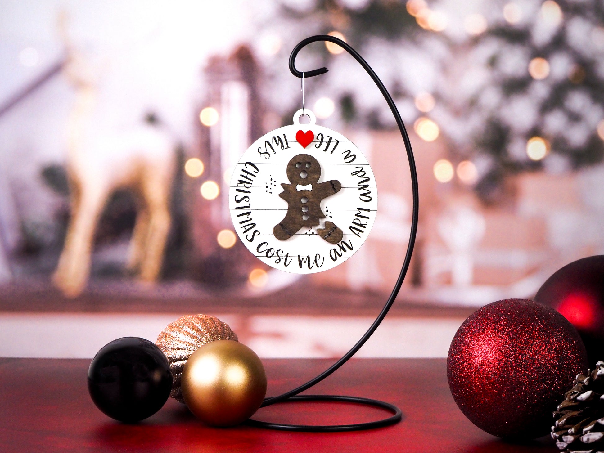 Hanging ornament featuring a distressed gingerbread man with a missing arm and leg, surrounded by the words, “This Christmas cost me an arm and a leg.” Displayed on a stand with a festive background.