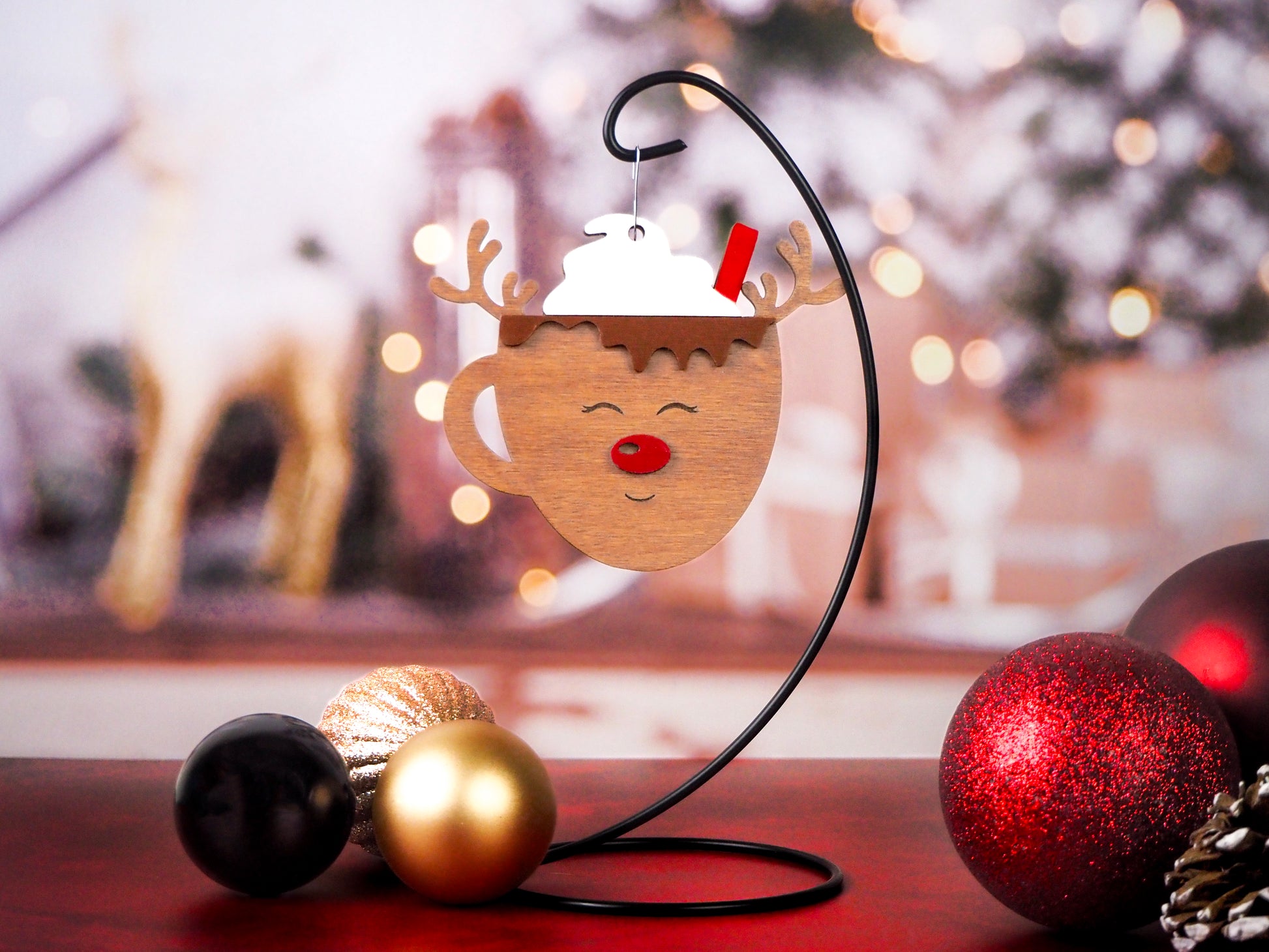 Hanging ornament in the shape of a reindeer mug with whipped cream and a red straw on top, complete with reindeer antlers and a red nose. Displayed on a stand with a festive background.