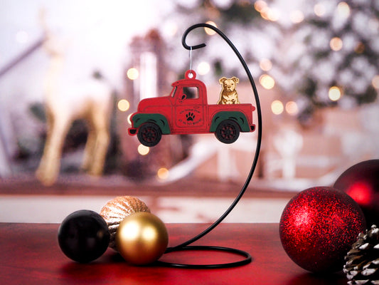 Ornament in the shape of a red vintage truck with a dog sitting in the back, hanging on a display stand with a festive holiday background.