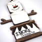 Close-up of a snowman ornament made of marshmallows, with twig-like arms and a wooden base that reads "S'MORE Christmas Fun."