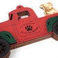 Close-up of a red truck ornament with green fenders, featuring a dog sitting in the truck bed. The door reads "Oh What Fun It Is To Ride" with a paw print design.