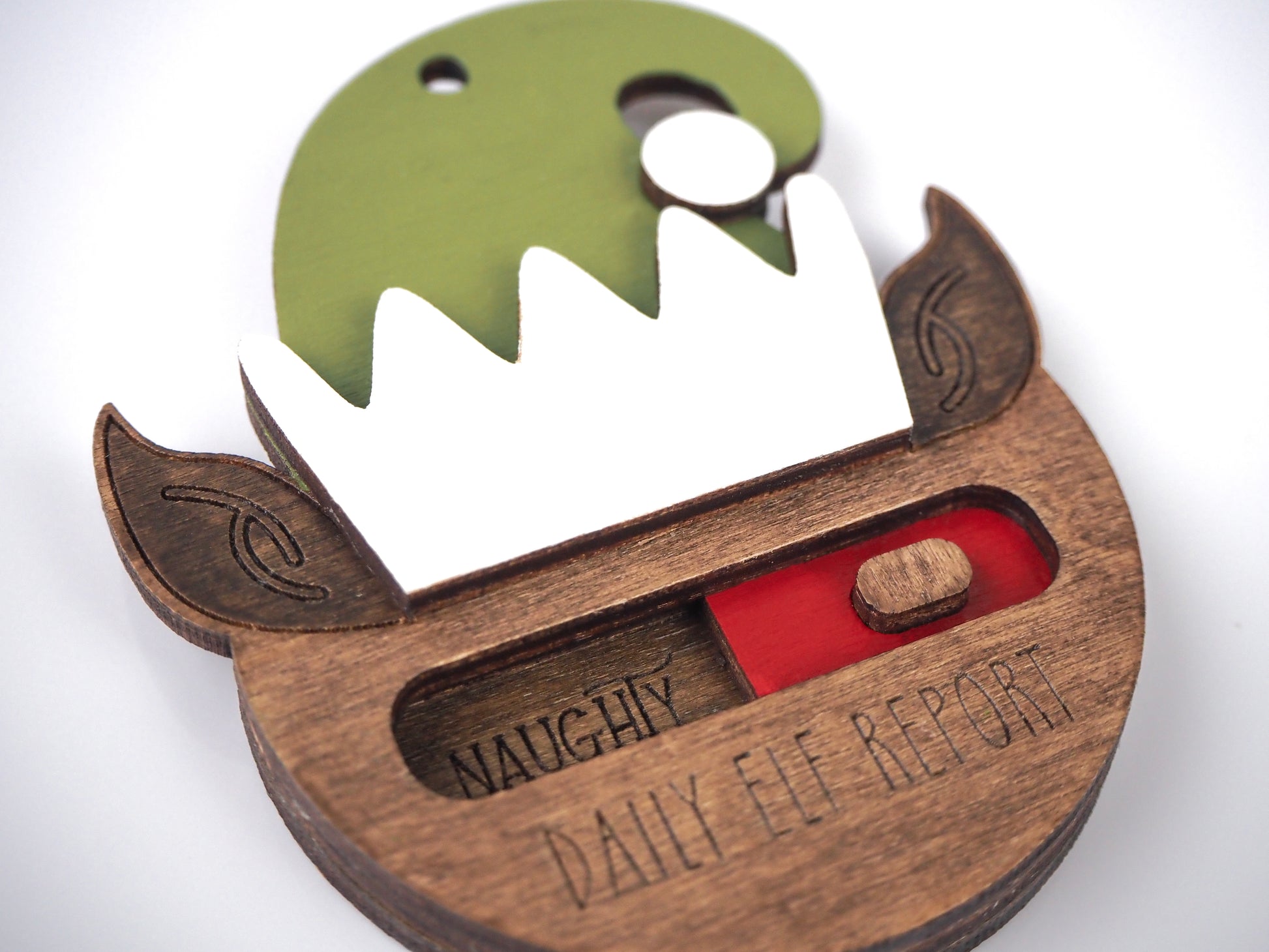 Close-up of the Daily Elf Report ornament, showcasing a green elf hat, brown elf ears, and a sliding indicator set to "Naughty." The ornament features engraved text reading "Daily Elf Report."