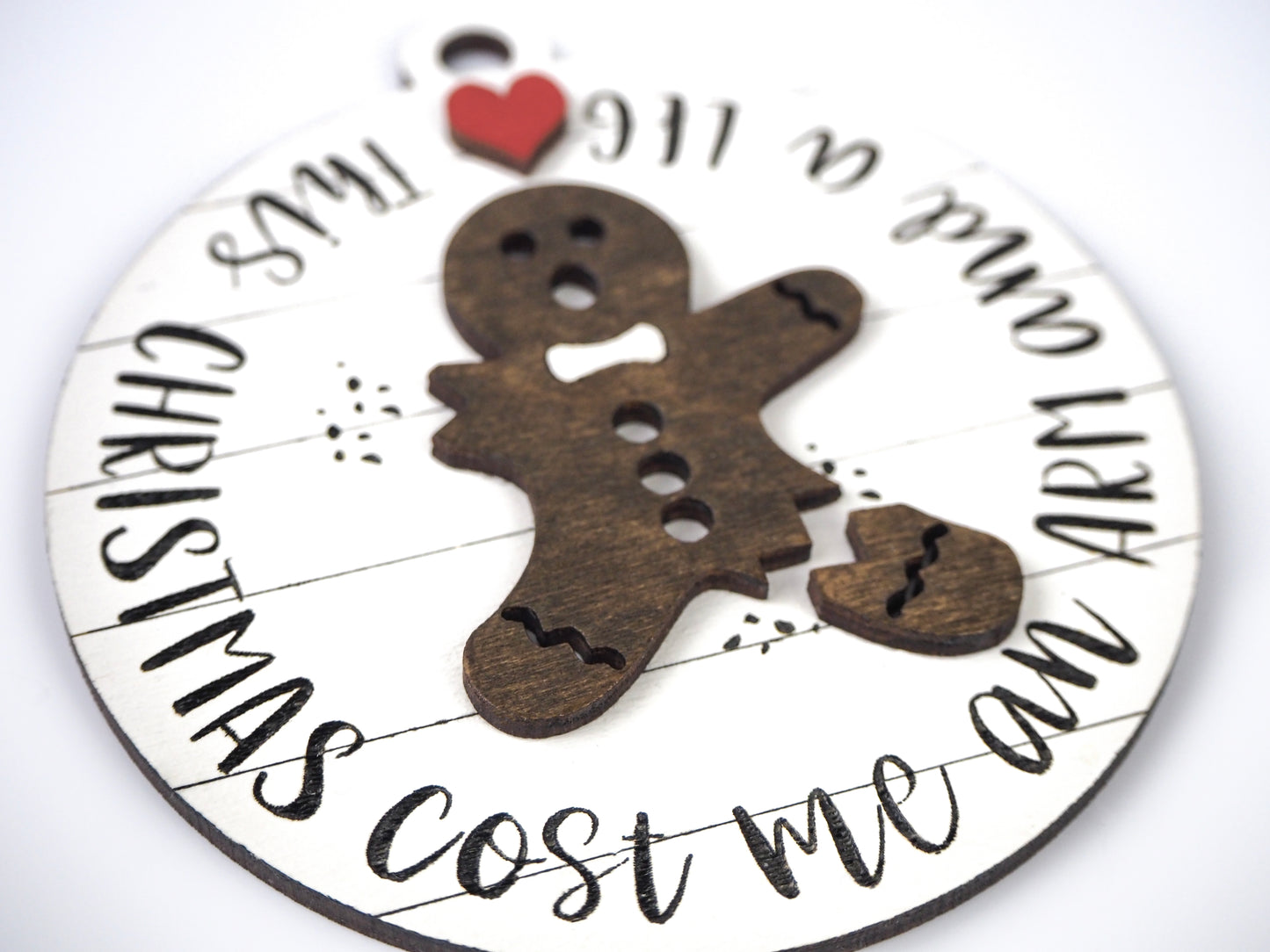 Close-up of a round ornament featuring a wooden gingerbread figure with a missing arm and leg. The surrounding text reads "Christmas Cost Me an Arm and a Leg," with a red heart detail at the top.