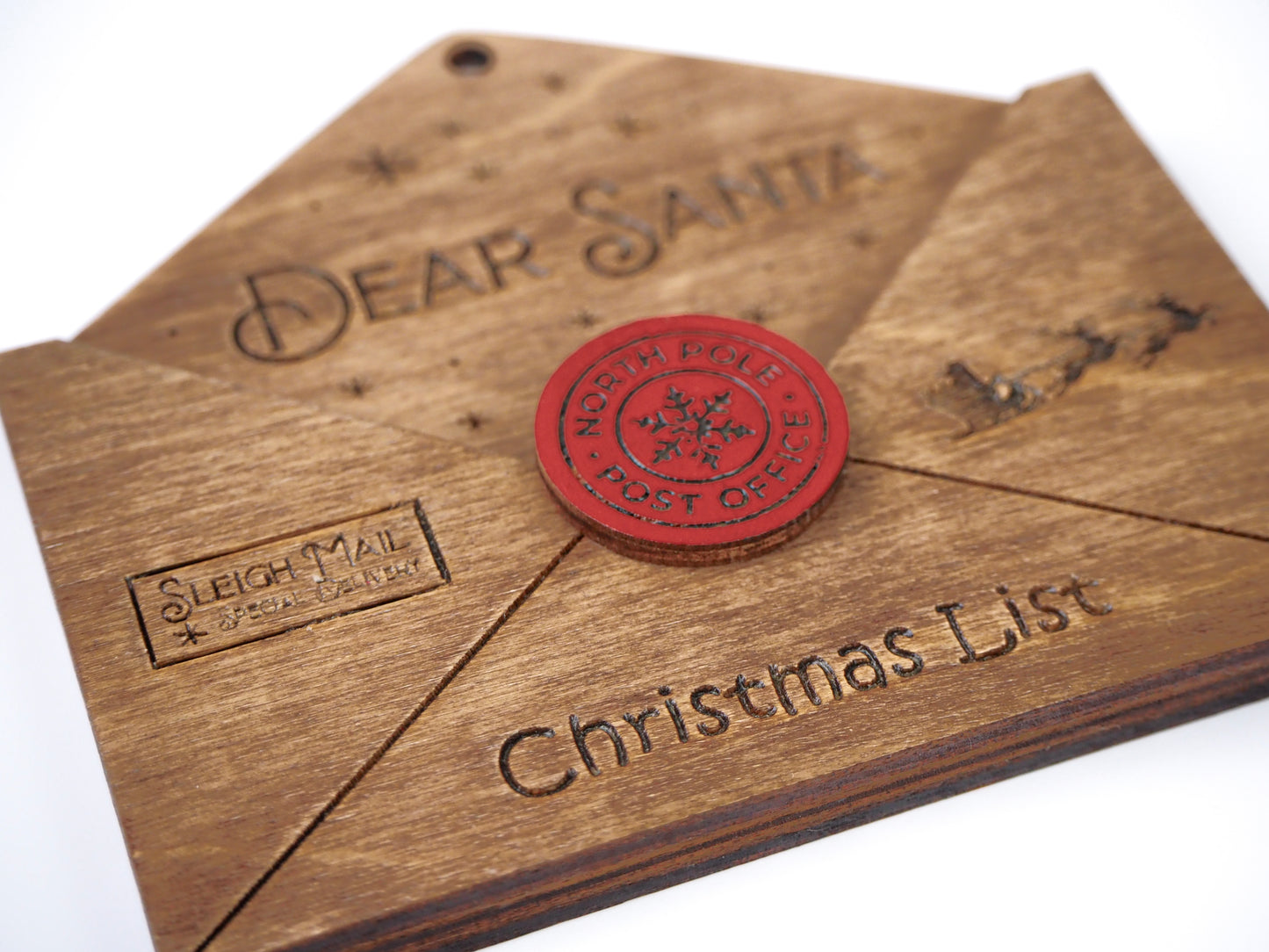 Close-up of a wooden "Dear Santa" envelope ornament with engraved details. Features a red "North Pole Post Office" seal, "Sleigh Mail" stamp, and "Christmas List" text on the bottom.