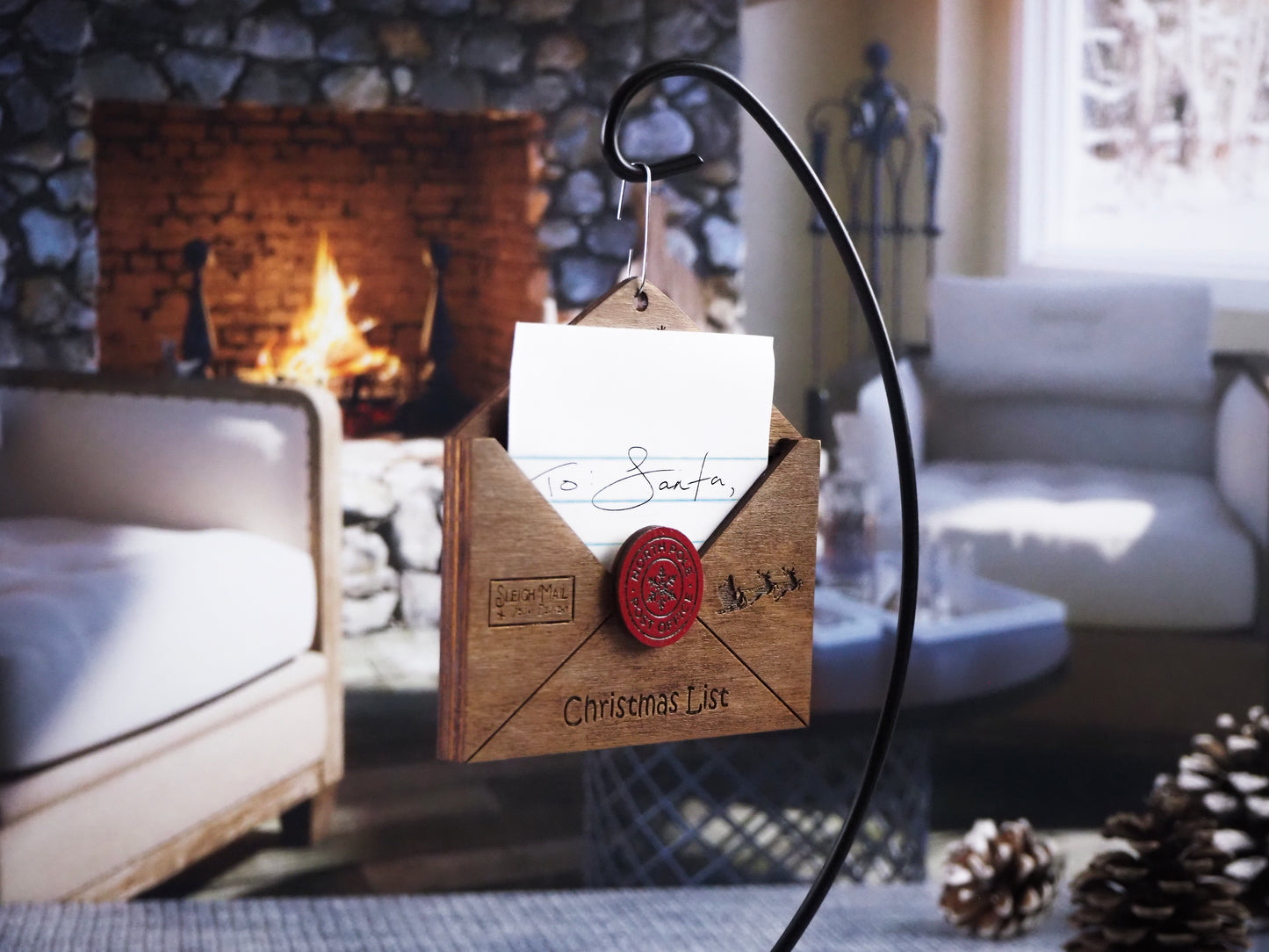 Wooden "Dear Santa" envelope ornament with a handwritten letter partially inside. Features a red "North Pole Post Office" seal, "Sleigh Mail" stamp, and "Christmas List" text, displayed in front of a cozy fireplace.