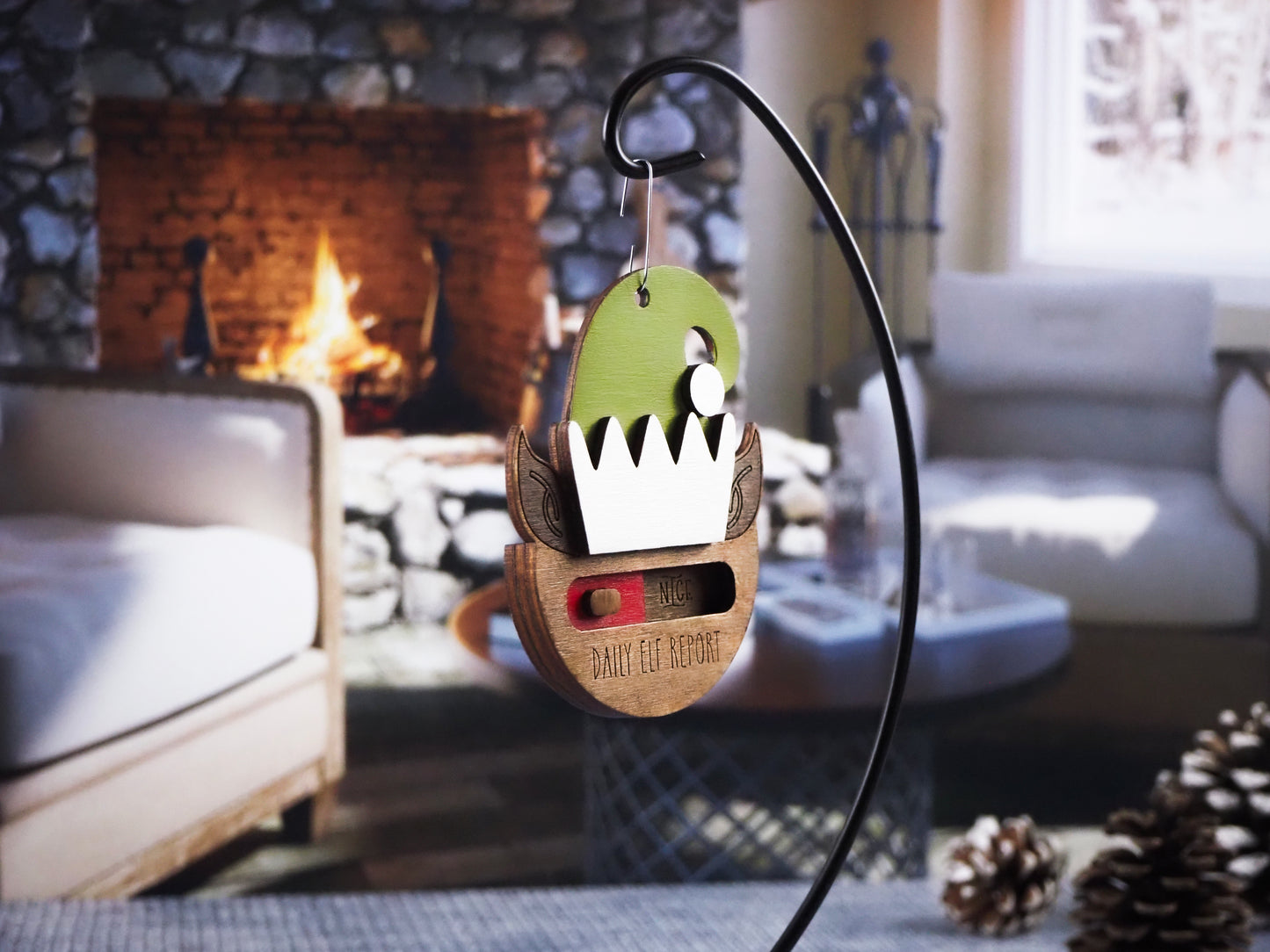 Wooden "Daily Elf Report" ornament with a green elf hat and a slider to choose between "Naughty" or "Nice." The ornament hangs in front of a cozy fireplace, creating a warm holiday atmosphere.