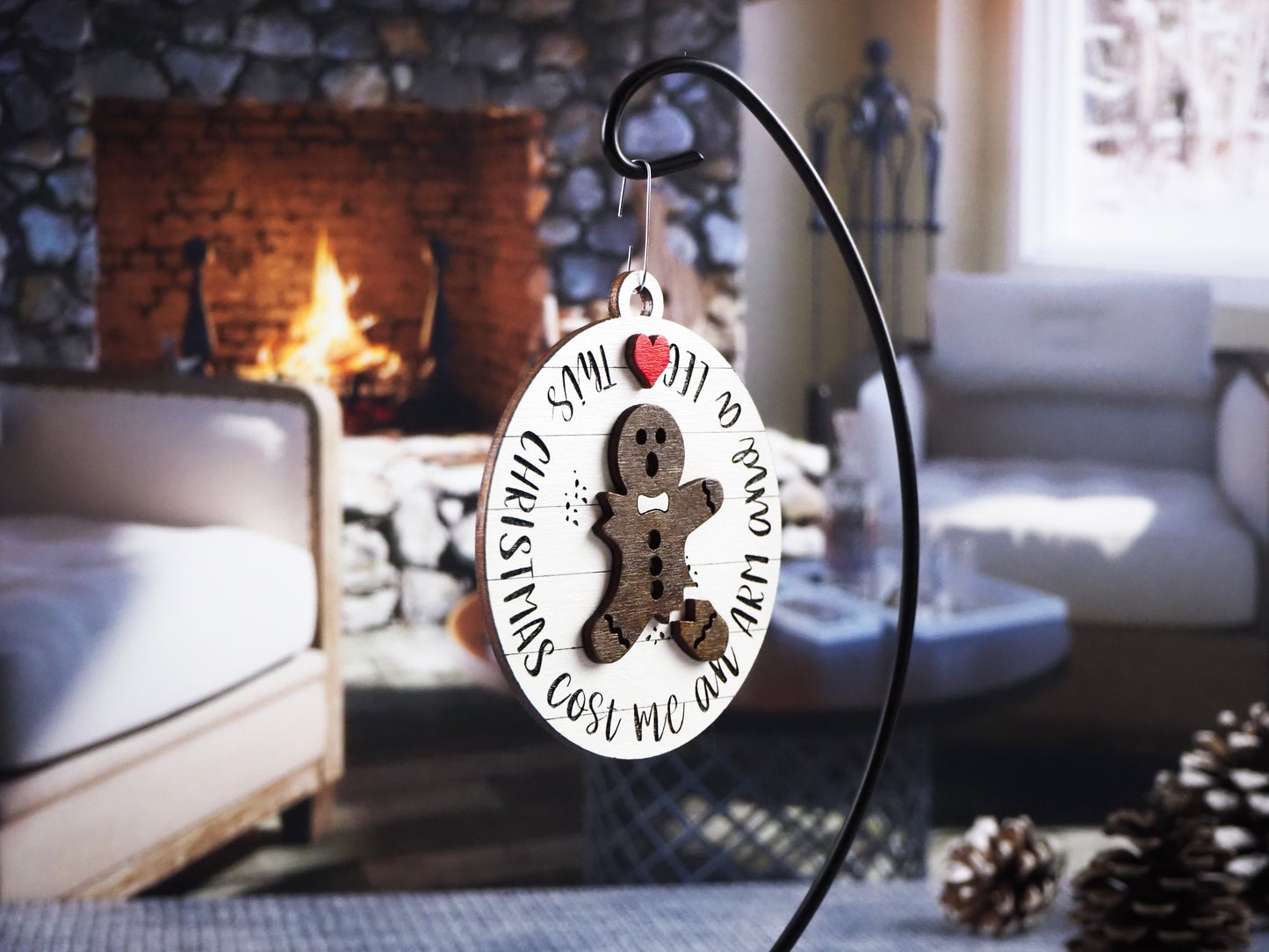 Round wooden ornament featuring a broken gingerbread man with a missing arm and leg. The words "Christmas Cost Me an Arm and a Leg" wrap around the design. The ornament hangs in front of a cozy fireplace.