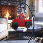 Wooden ornament of a red vintage truck with green fenders, featuring a dog sitting in the back. The truck displays the text "Oh What Fun It Is to Ride!" The ornament hangs in front of a cozy fireplace setting.