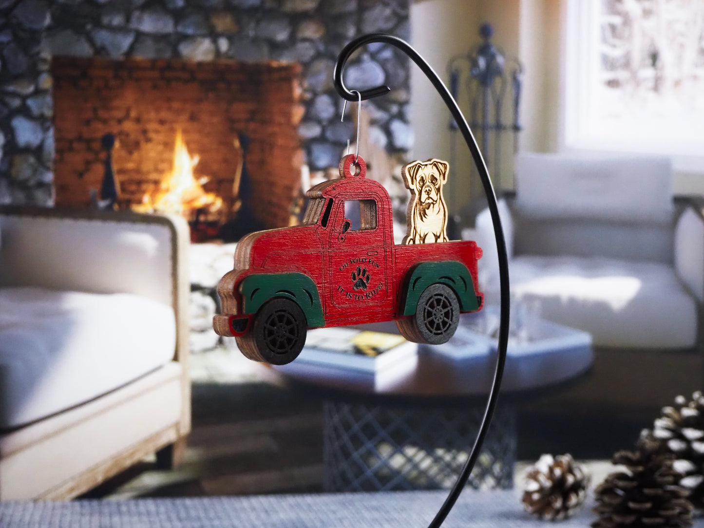 Wooden ornament of a red vintage truck with green fenders, featuring a dog sitting in the back. The truck displays the text "Oh What Fun It Is to Ride!" The ornament hangs in front of a cozy fireplace setting.