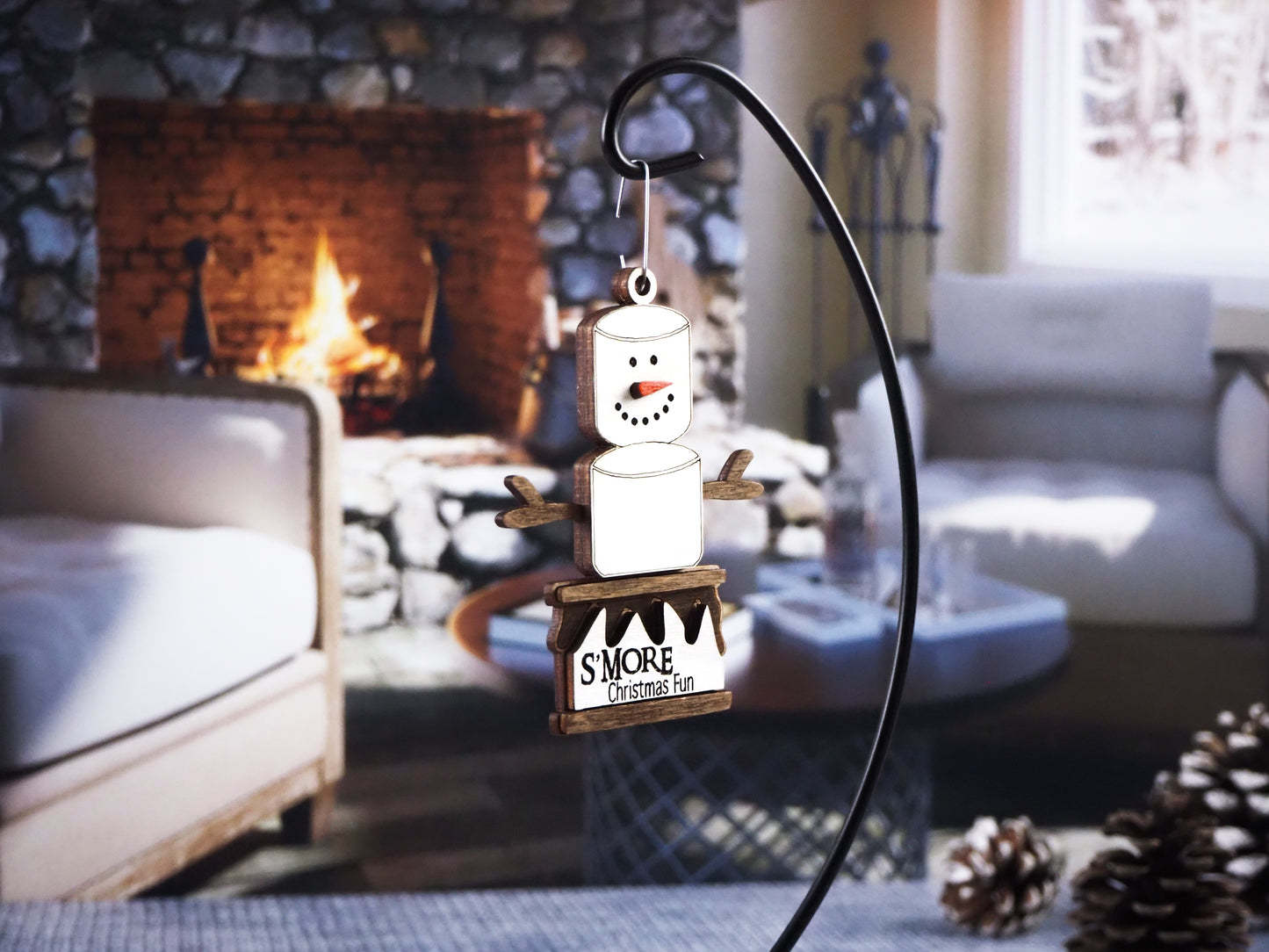 Wooden ornament shaped like a snowman made of marshmallows, with twig arms and sitting on a graham cracker base. The base has text that reads "S'more Christmas Fun." The ornament is displayed in front of a warm fireplace setting.