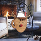 Wooden ornament shaped like a reindeer mug with antlers, whipped cream, and a straw on top. The reindeer mug features a red nose and a smiling face. The ornament is displayed in front of a warm fireplace setting.