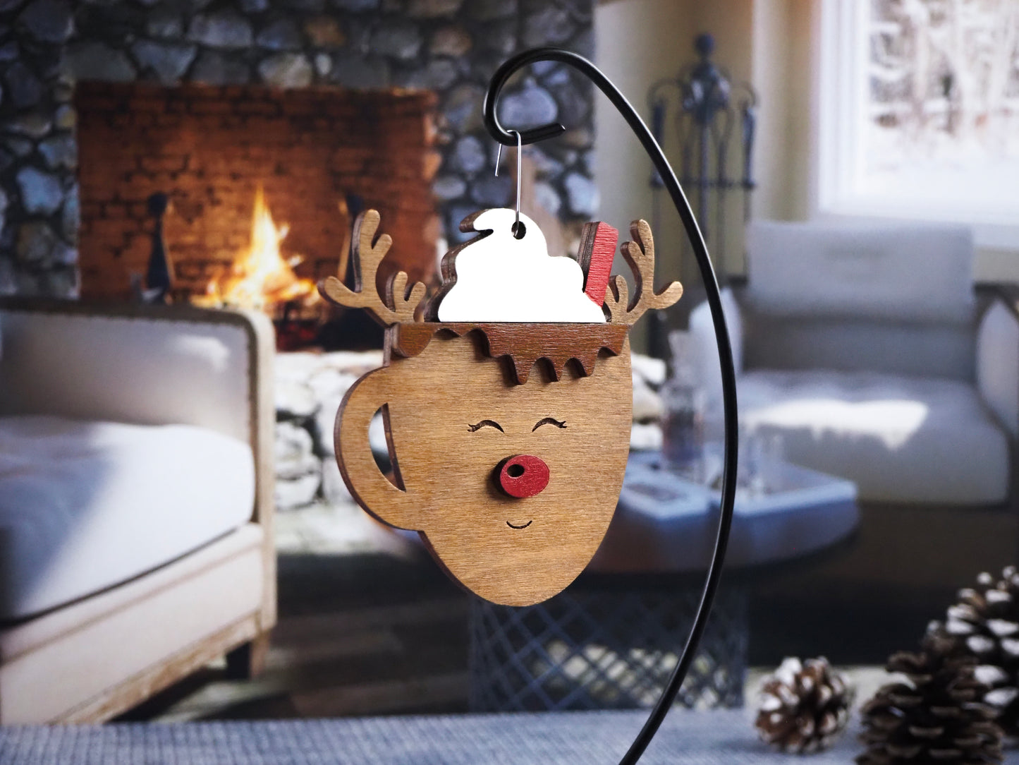 Wooden ornament shaped like a reindeer mug with antlers, whipped cream, and a straw on top. The reindeer mug features a red nose and a smiling face. The ornament is displayed in front of a warm fireplace setting.