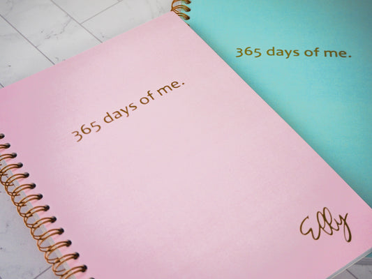 Two spiral-bound notebooks with "365 days of me." written in gold on the cover. The front notebook is pink with the personalized name "Elly" in the corner, while the back notebook is turquoise.