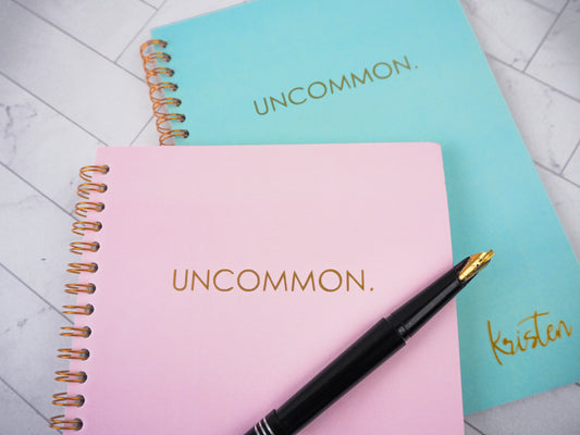 Two spiral-bound notebooks with pink and turquoise covers, both featuring the word "UNCOMMON" in gold lettering. The turquoise notebook also shows the personalized name "Kristen" in gold. A black pen rests on the pink notebook.